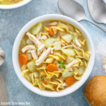 Chicken noodle soup in a white bowl sitting on a blue counter with spoons and a piece of bread.
