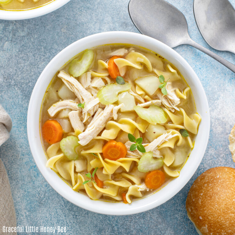 Chicken noodle soup in a white bowl sitting on a blue counter with spoons and a piece of bread.