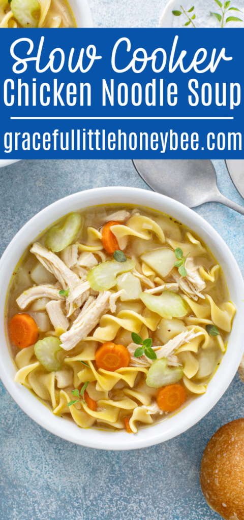 Chicken noodle soup in a white bowl sitting on a blue counter with spoons and a piece of bread.