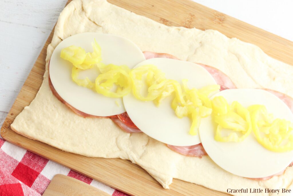 Crescent roll dough lying flat with ham, cheese and banana peppers layered on top.