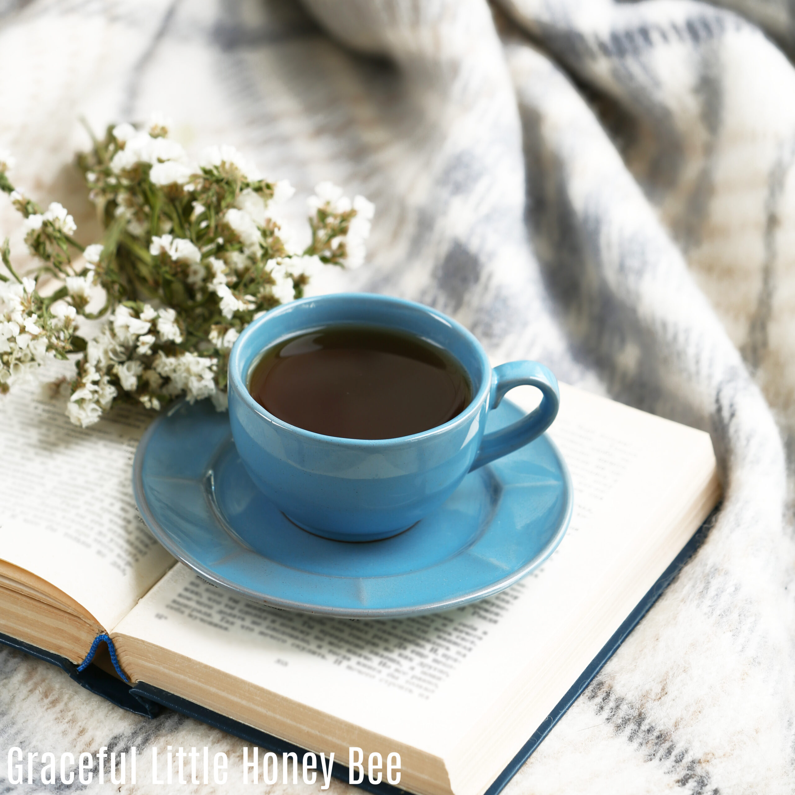 An open book with a blue coffee cup and saucer sitting on top along with a bouquet of baby's breath.