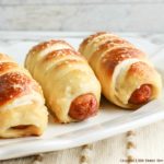 Three golden brown pretzel dogs sitting in a row on a white plate.