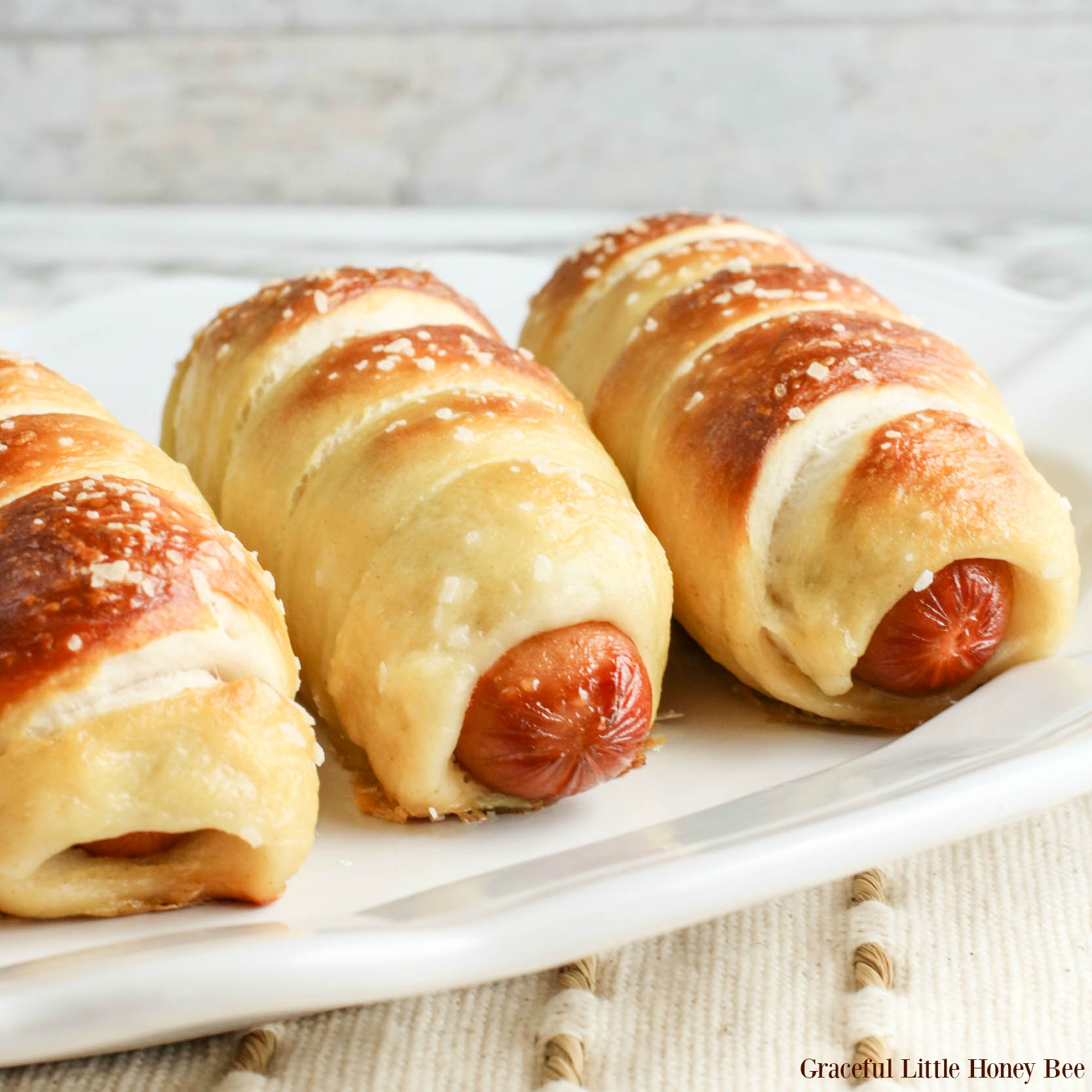 Three golden brown pretzel dogs sitting in a row on a white plate.