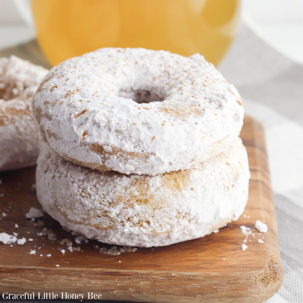 Two Apple Cider Donuts stacked on a wooden cutting board with a glass of apple cider in the background.