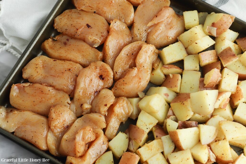 Raw seasoned chicken and raw seasoned potatoes on a baking sheet.