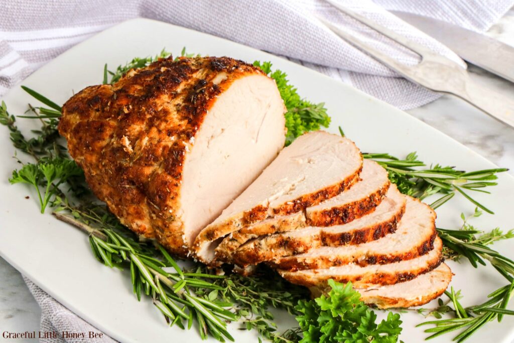 Air Fryer Turkey Breast, sliced on a white platter surrounded by fresh herbs.