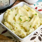 Fluffy mashed potatoes in a white, square serving bowl with a pat of butter and fresh parley sprinkled on top.