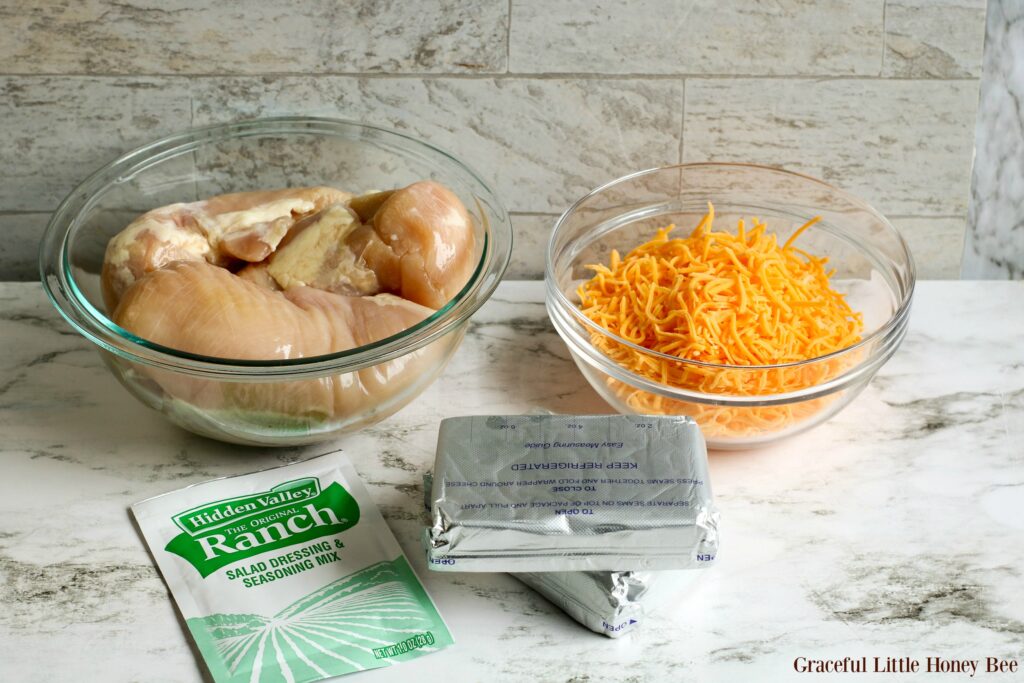 Ingredients sitting on a marble countertop including, raw chicken breast, shredded cheddar cheese, cream cheese and ranch dressing powder.