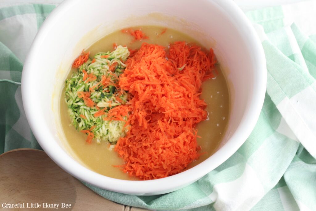 Muffin batter in a white bowl, with shredded zucchini and carrot on top before mixing.