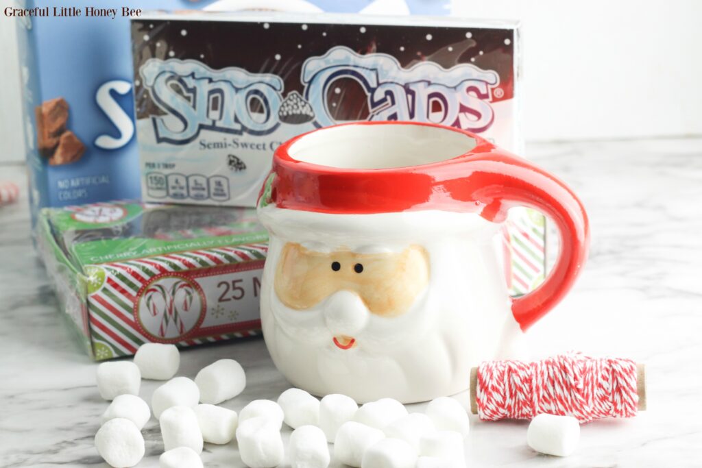 Empty Santa Mugi with marshmallows, candy canes, chocolate candy and hot choclate sitting next to it.