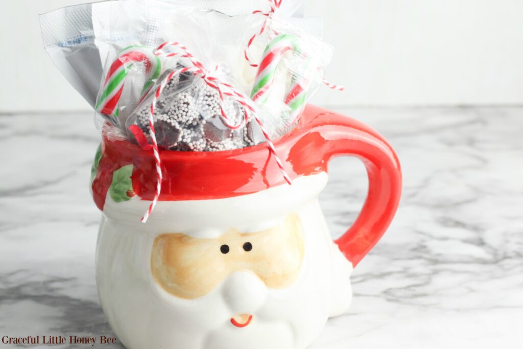 Santa Mug filled with hot chocolate packets, candy canes, marshmallows and chocolate candy sitting on a marble countertop.