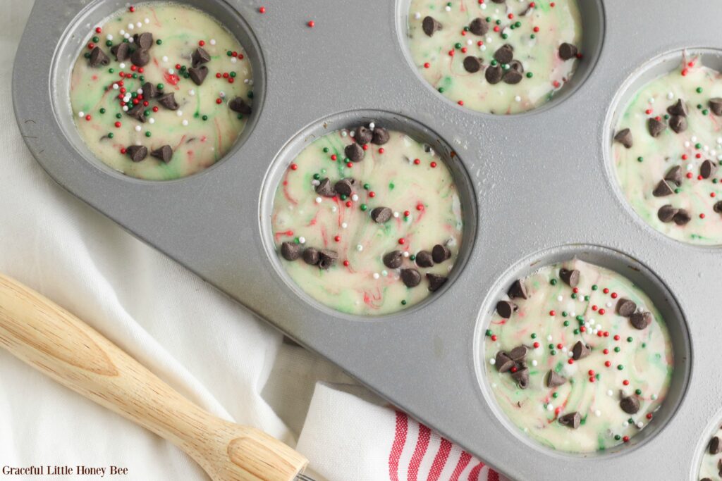 Muffin batter sprinkled with chocolate chips and sprinkles in a muffin tin.