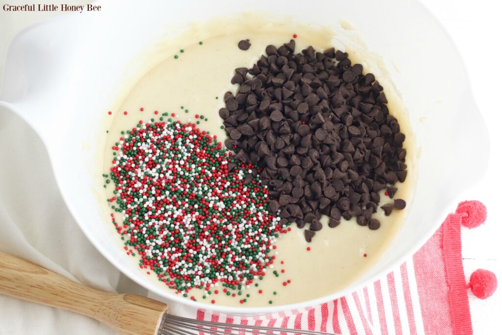 Muffin batter in a white mixing bowl with a pile of sprinkles and chocolate chips on top.