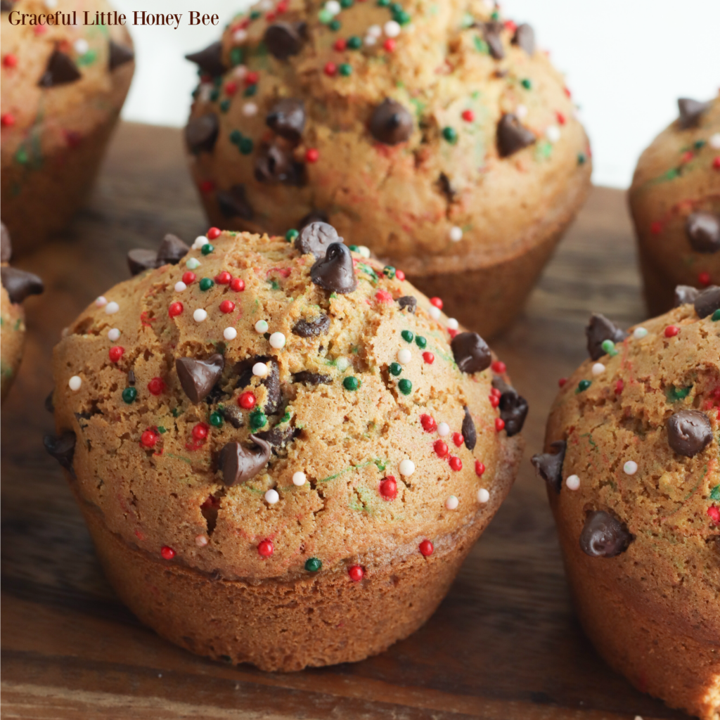 Chocolate chip muffins sprinkled with red, white and green sprinkles sitting on a wooden cutting board.