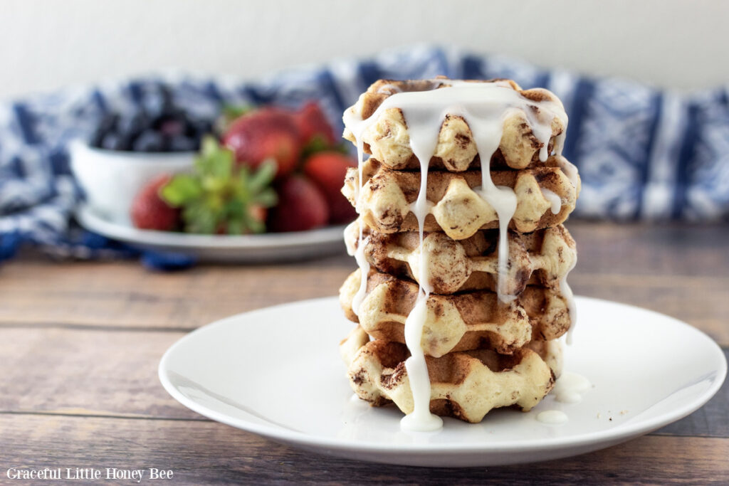 A stack of cinnamon roll waffles on a white plate with fresh fruit in the background.