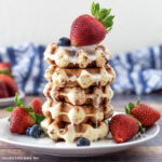 Cinnamon Roll Waffles stacked on a plate drizzled with glaze and topped with fresh fruit.