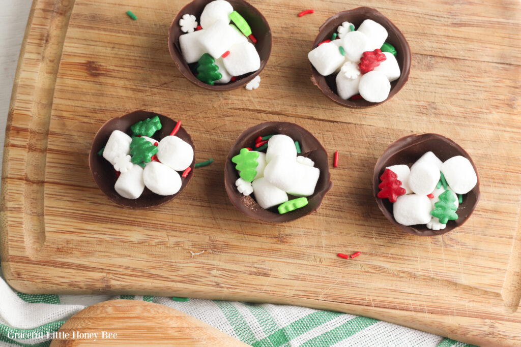 Half of the hardened chocolate filled with hot cocoa mix, mini marshmallows and holiday sprinkles sitting on a wooden cutting board.