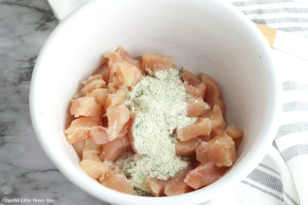 Raw chicken pieces in white bowl with ranch seasoning on top.