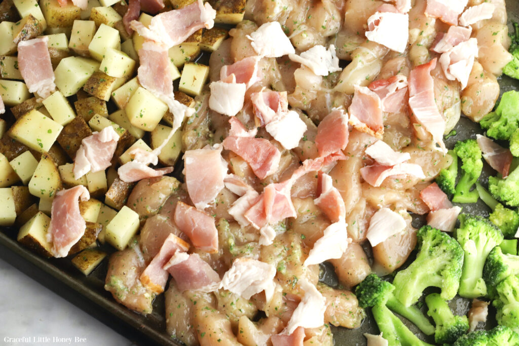 Potatoes, raw chicken, raw bacon and broccoli on a sheet pan before baking.