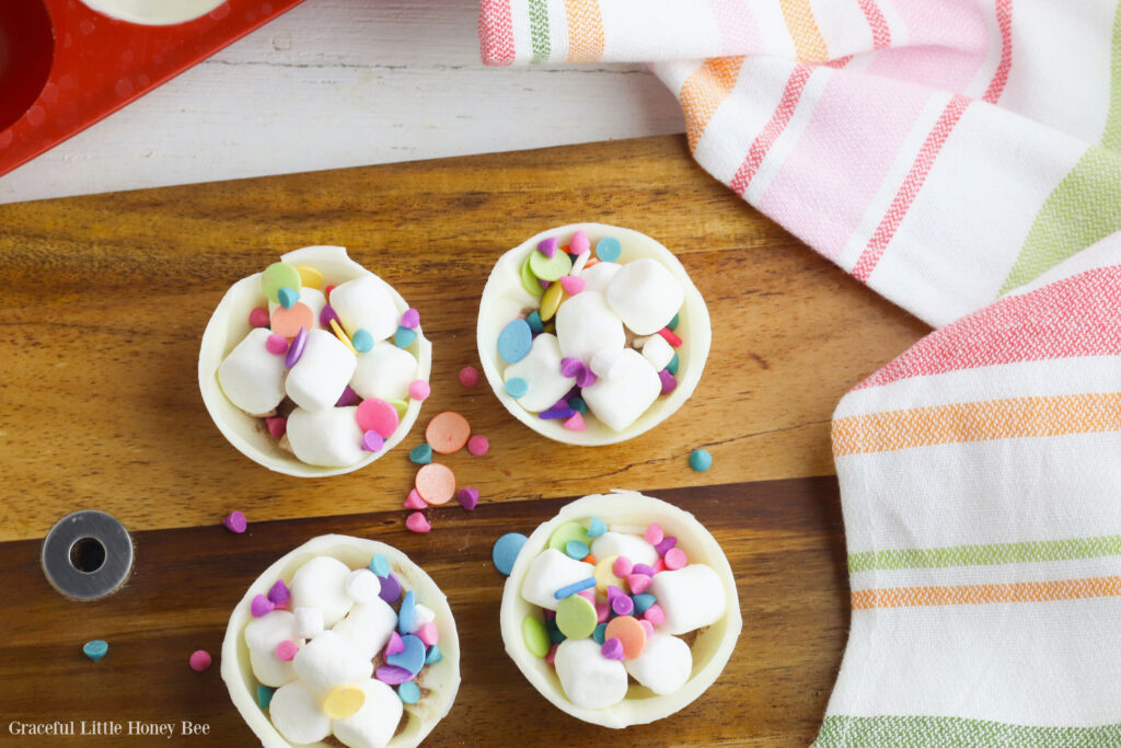 Four half shells of a hot chocolate bomb filled with instant hot chocolate, mini marshmallows and rainbow sprinkles sitting on a wooden cutting board.