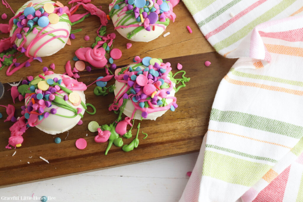 White hot chocolate bombs covered with rainbow icing and sprinkles on a brown cutting board.