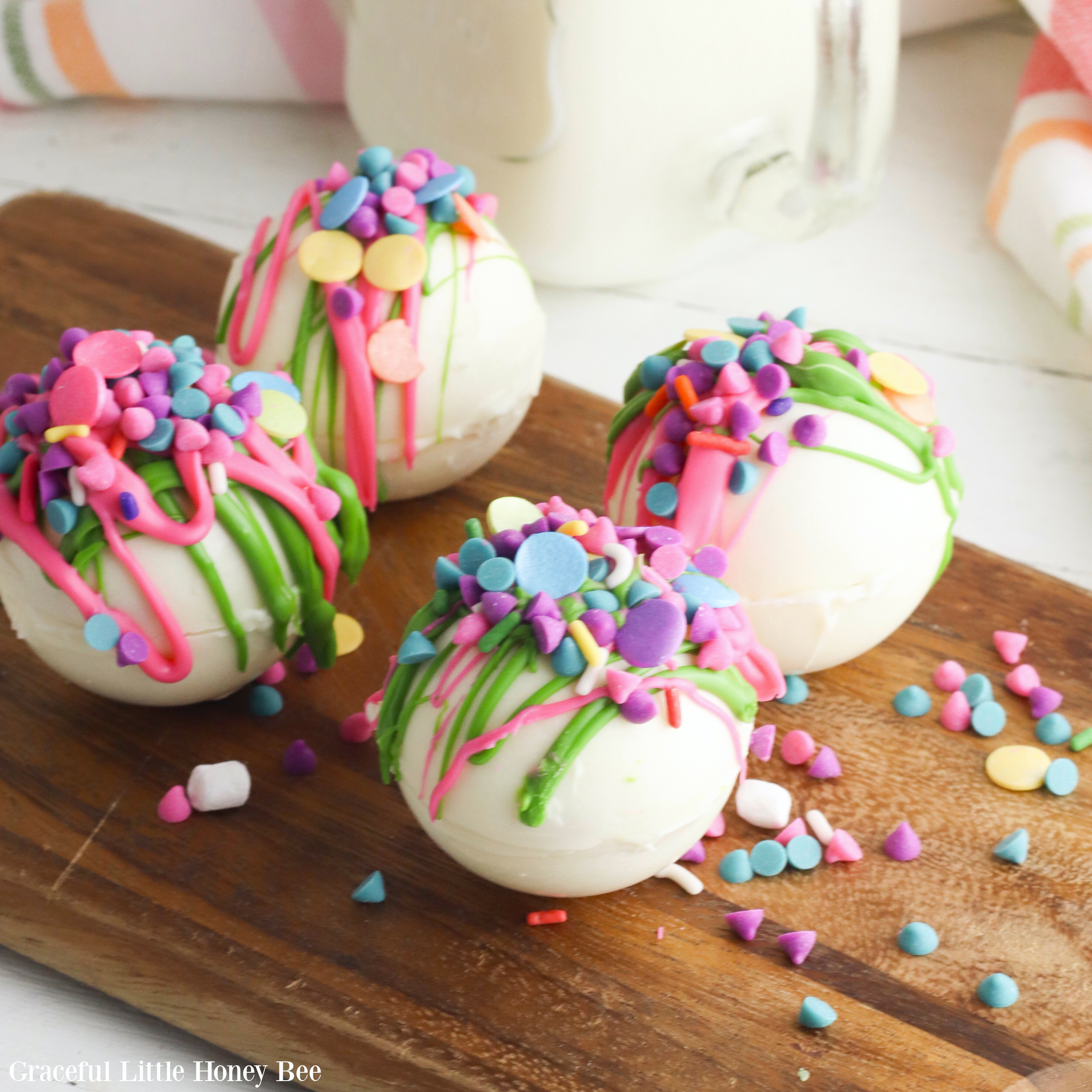 White hot chocolate bombs covered with rainbow icing and sprinkles on a brown cutting board.