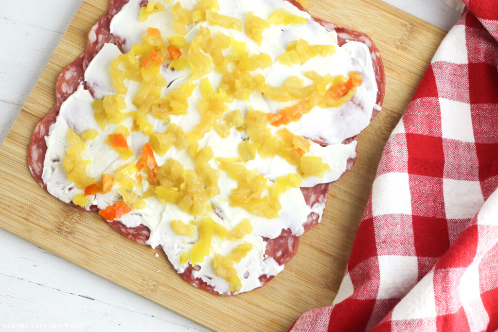 A layer of salami with cream cheese and pickled peppers on top sitting on a wooden cutting board.