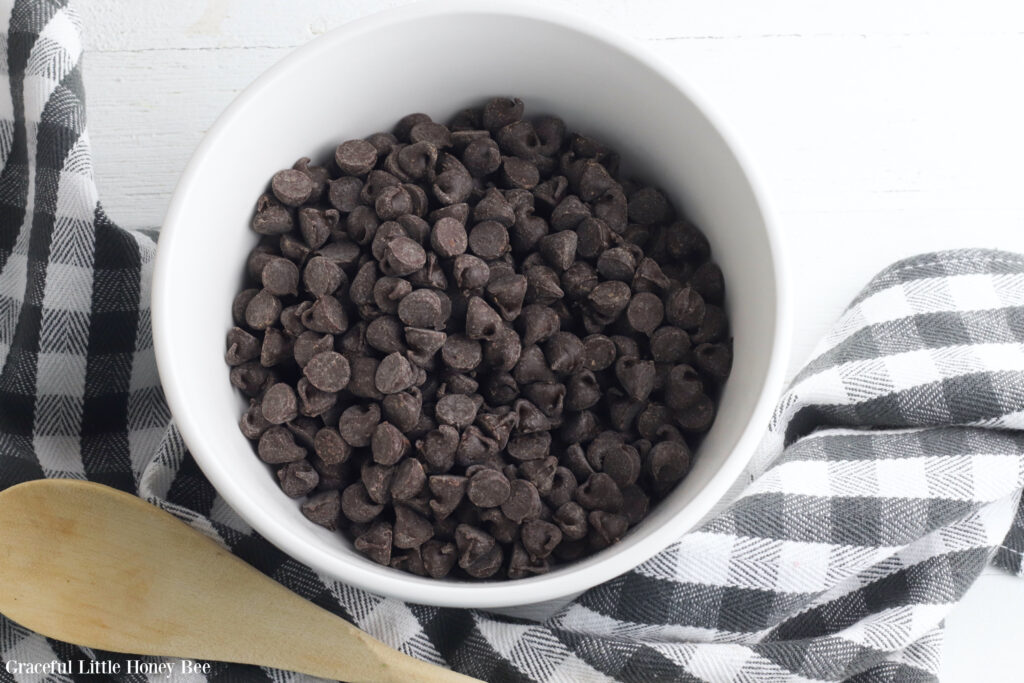 Chocolate chips in a small white bowl with a wooden spoon laying next too it.