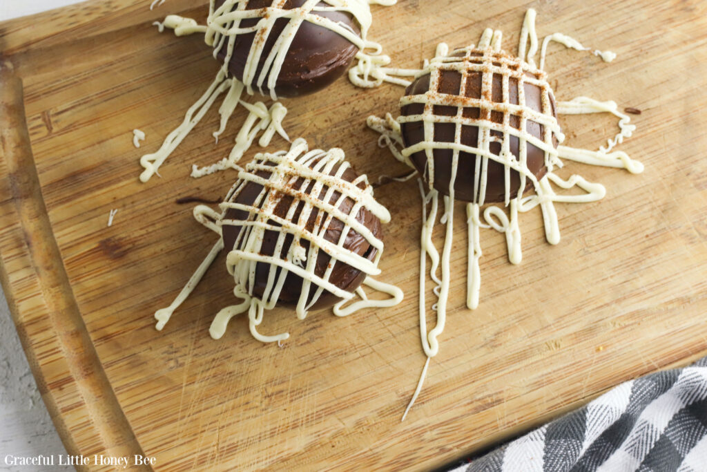 A set of three Mocha Hot Chocolate Bombs sitting on a wooden cutting board with white chocolate icing drizzled over the tops.