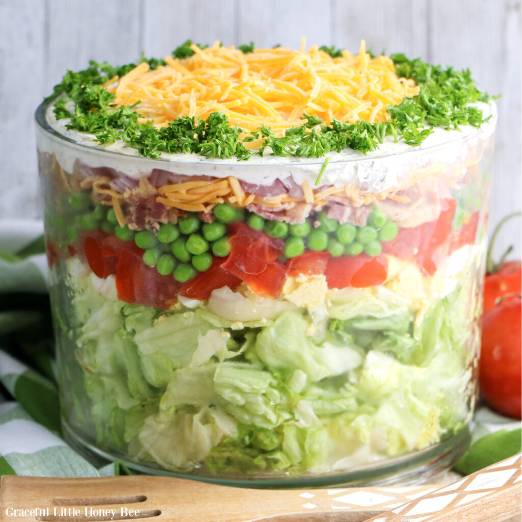 7-Layer Salad in a clear glass trifle bowl sitting on a marble counter with a wooden spoon sitting next to it.