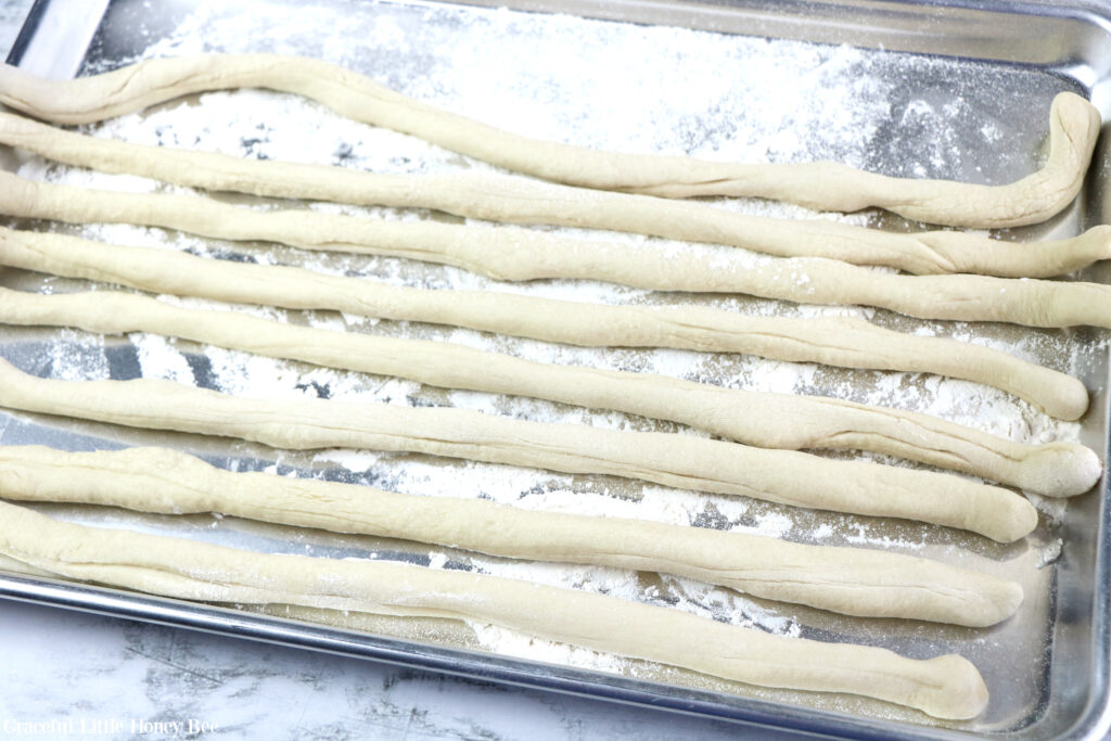 Eight pieces of dough rolled into long pieces, sitting on a floured countertop.