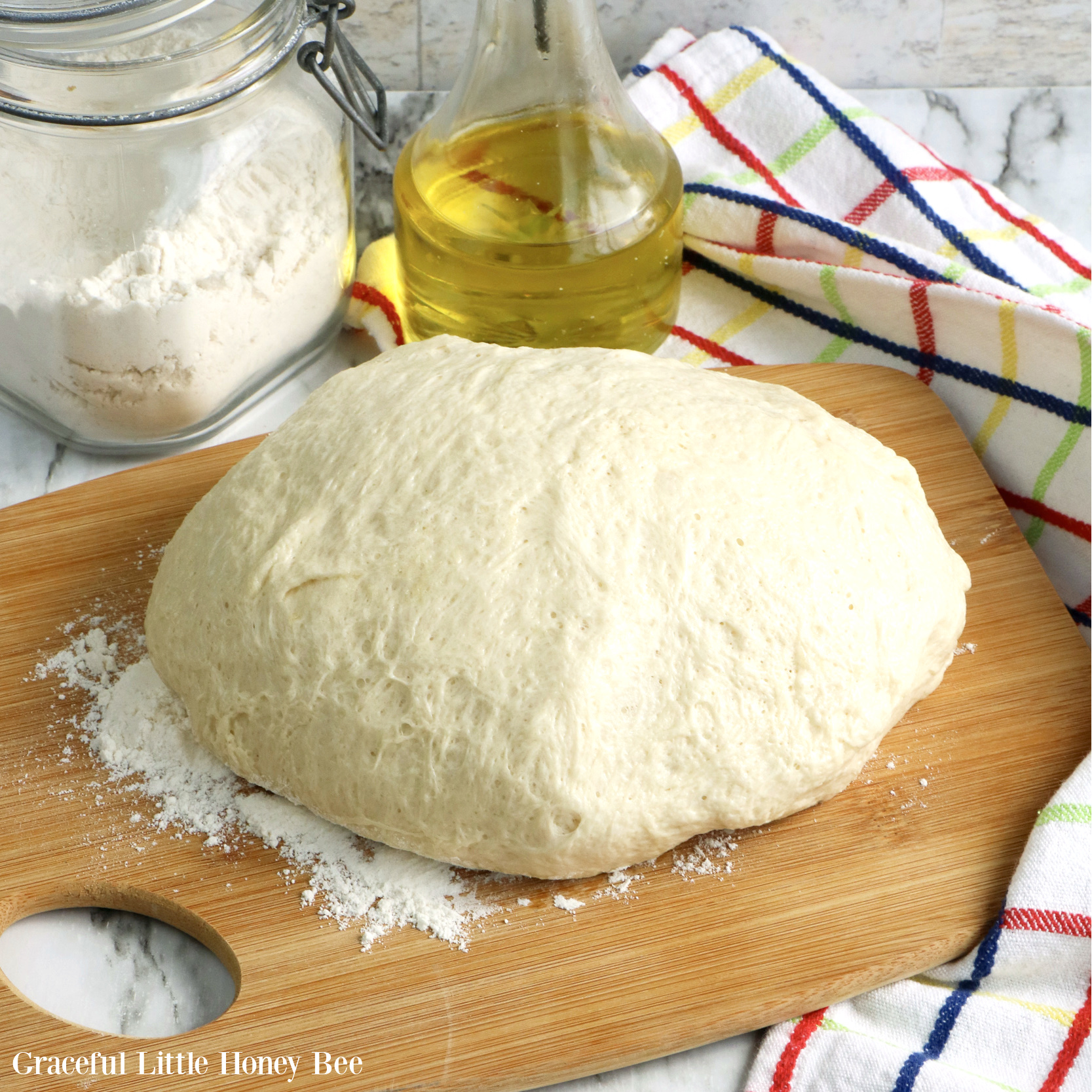 Bread dough sitting on a wooden cutting board with a bottle of oil and flour behind it.