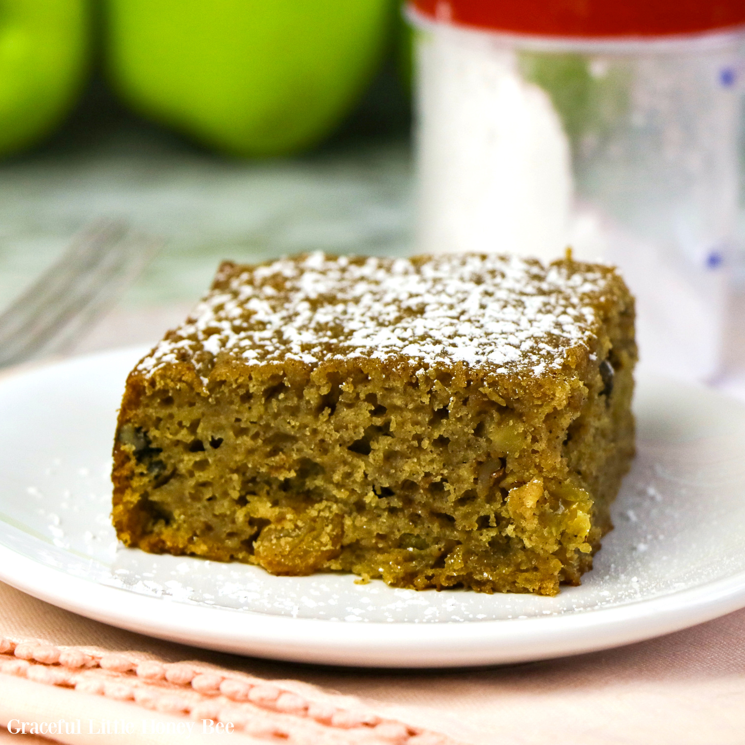 A slice of Applesauce topped with powdered sugar sitting on a white plate with green apples in the background.