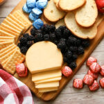 An assortment of red, white and blue finger food arranged neatly on a cutting board including strawberries, blueberries, crackers, cheese and chocolate.