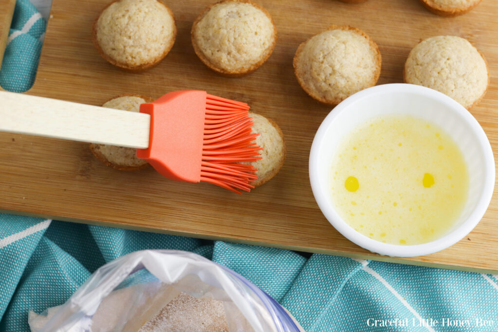 Mini muffins sitting on a wooden cutting board, being brushed with melted butter.