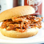 Pulled Pork Sandwhich sitting a on white plate with a bottle of Dr. Pepper in the background.