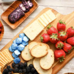 An assortment of red, white and blue finger food arranged neatly on a cutting board including strawberries, blueberries, crackers, cheese and chocolate.