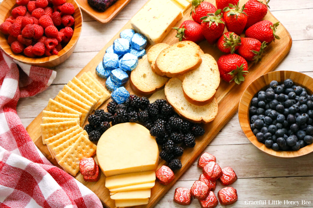 An assortment of red, white and blue finger food arranged neatly on a cutting board including strawberries, blueberries, crackers, cheese and chocolate.