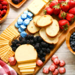 An assortment of red, white and blue finger food arranged neatly on a cutting board including strawberries, blueberries, crackers, cheese and chocolate.