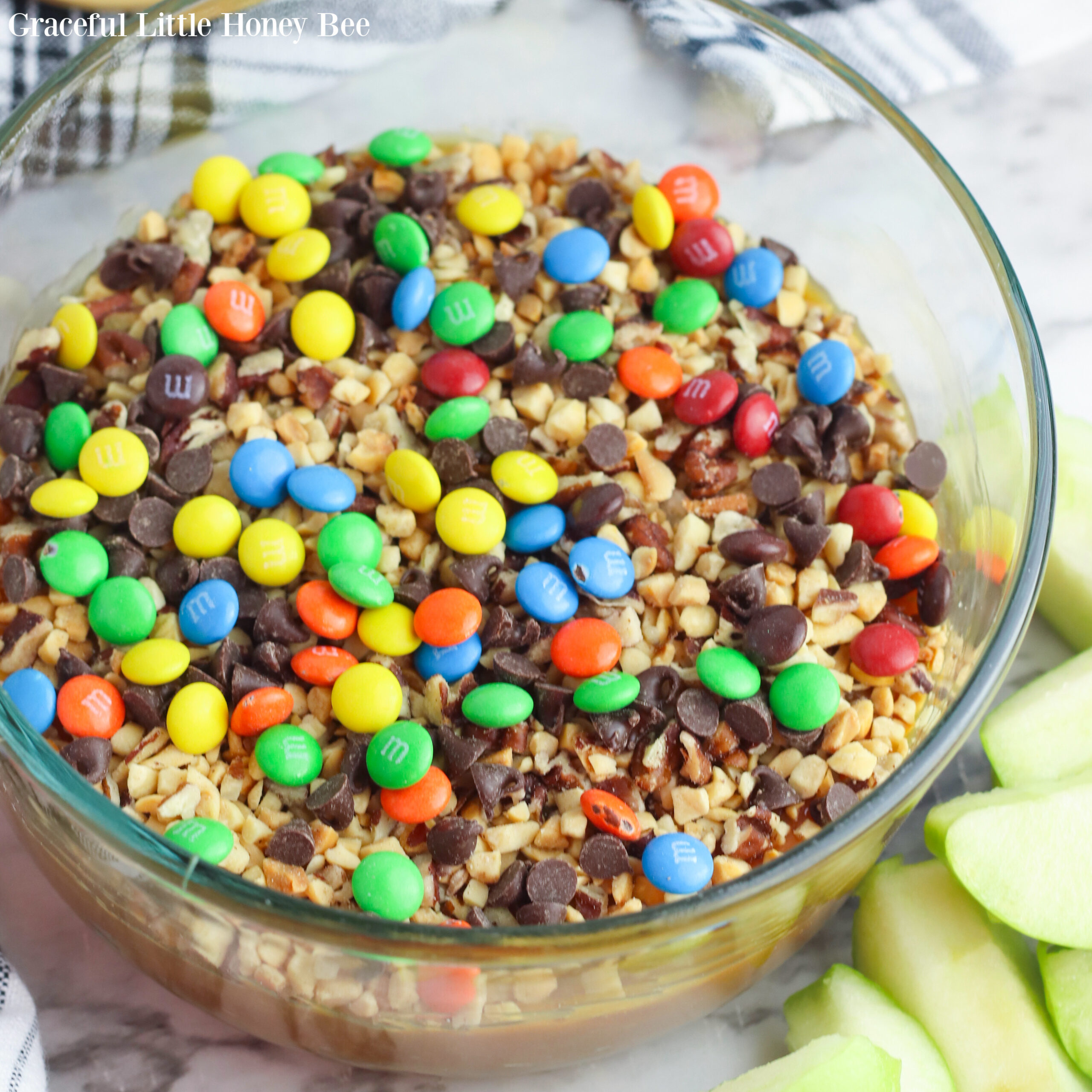 Caramel Apple dip with Peanuts and Chocolate Candy in a clear glass bowl with green apple slices sitting next to it.