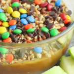 Caramel Apple dip with Peanuts and Chocolate Candy in a clear glass bowl with green apple slices sitting next to it.