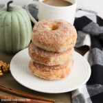 Three pumpkin spice donuts stacked on a white plate with a cup of coffee and a green pumpkin in the background.