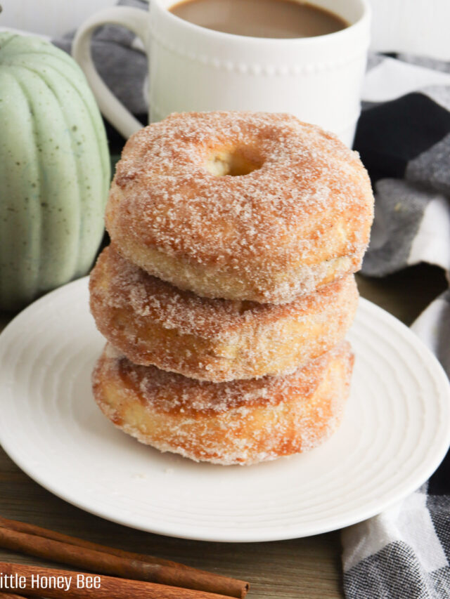 Three pumpkin spice donuts stacked on a white plate with a cup of coffee and a green pumpkin in the background.