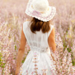 A woman standing in a flower field wearing a white lace dress and dainty white hat.