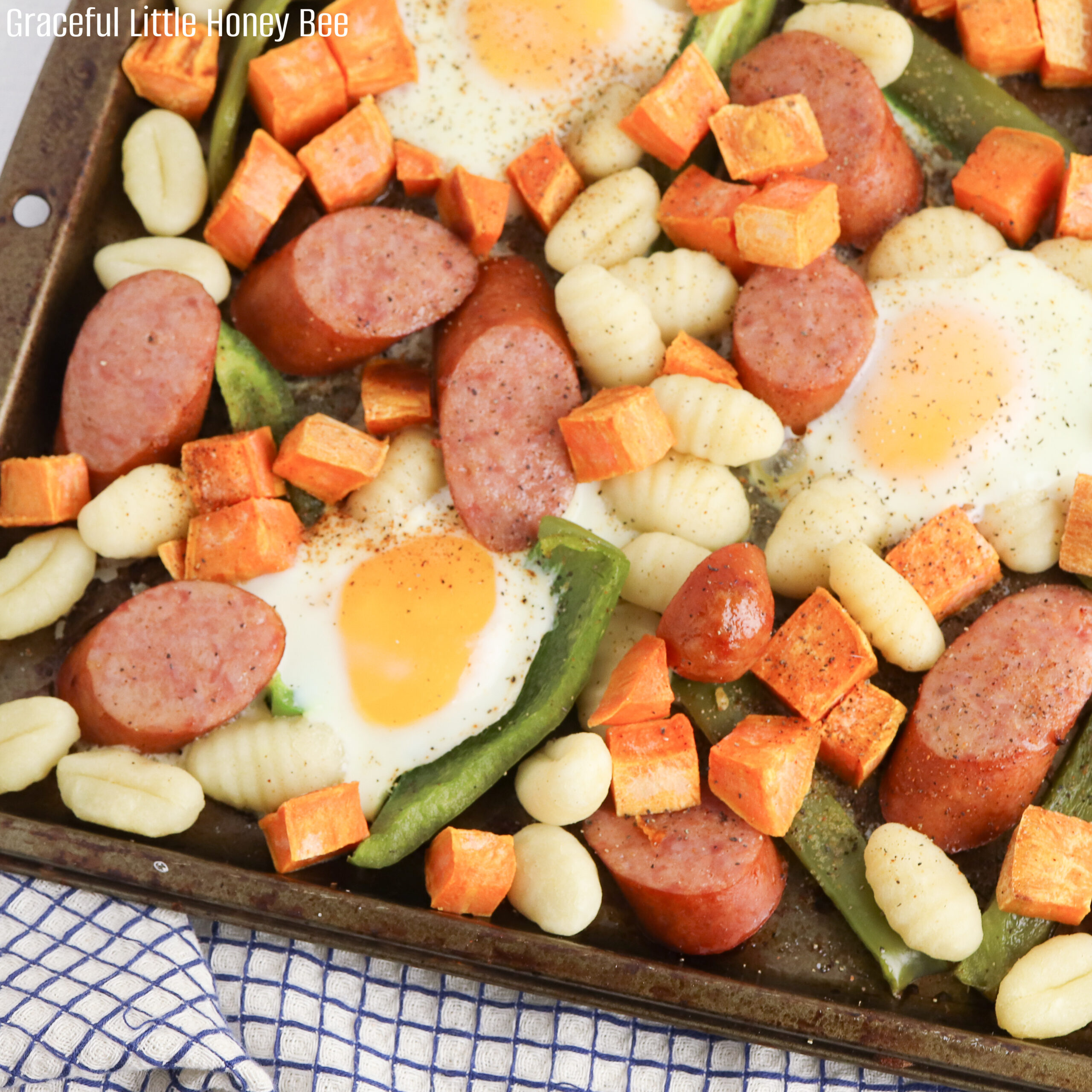 A close up of sliced sausage, diced sweet potatoes, gnocchi, bell pepper and eggs on a sheet pan.