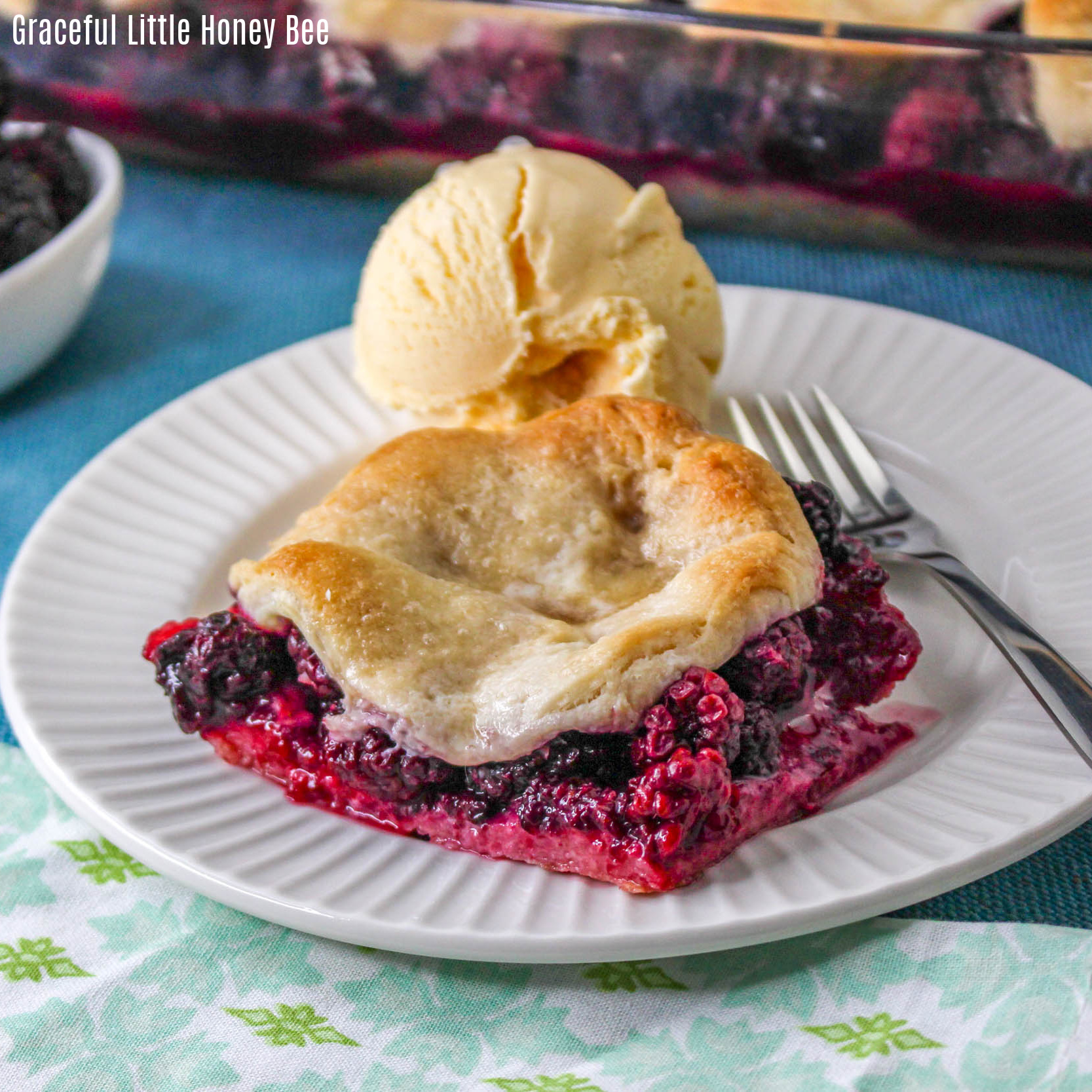 A Serving of golden brown blackberry cobbler on a round white plate with a large scoop of ice cream next to it.