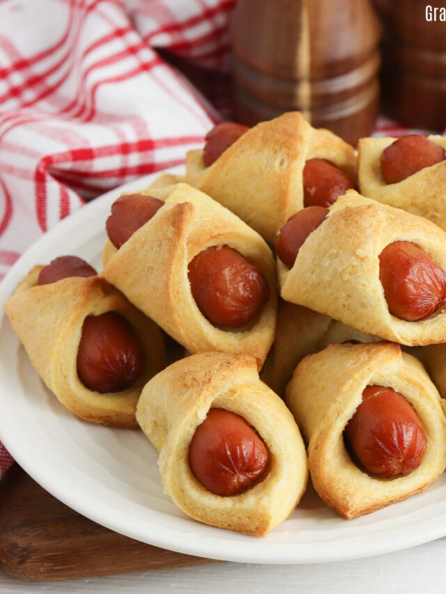 Corn dog pigs in a blanker sitting in a pile on a white plate.