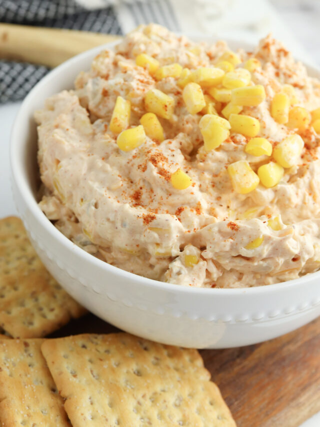 A close up view of corn dip with corn kernels on top for garnish in a white bowl.