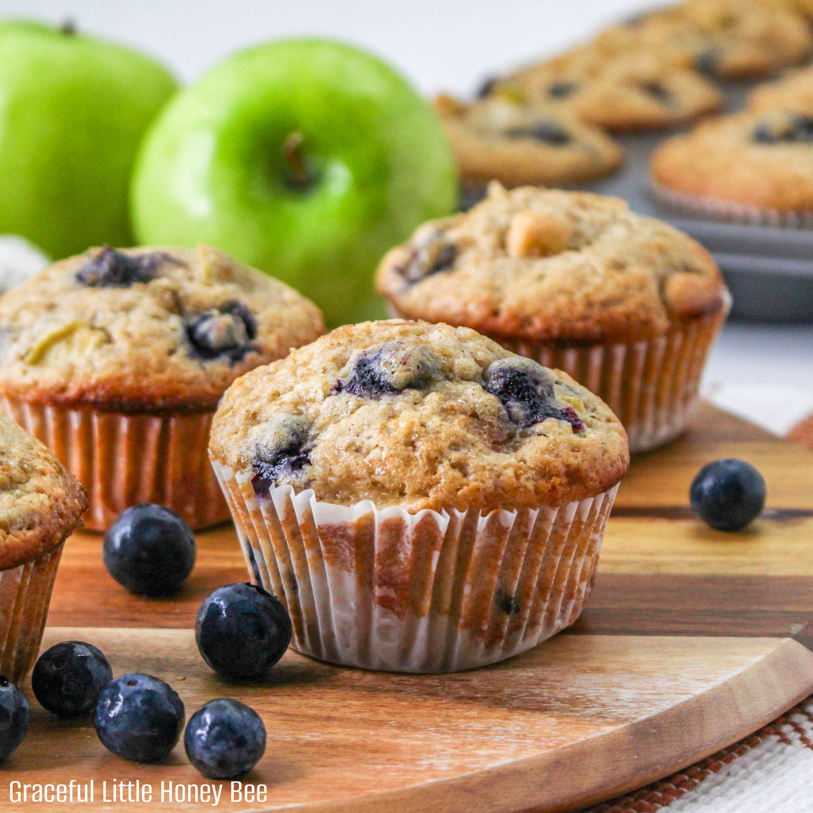 A close up view of blueberry apple muffins sitting on a wooden cutting board with a green apple in the background.