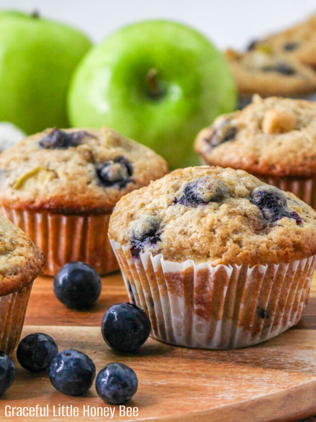 A close up view of blueberry apple muffins sitting on a wooden cutting board with a green apple in the background.